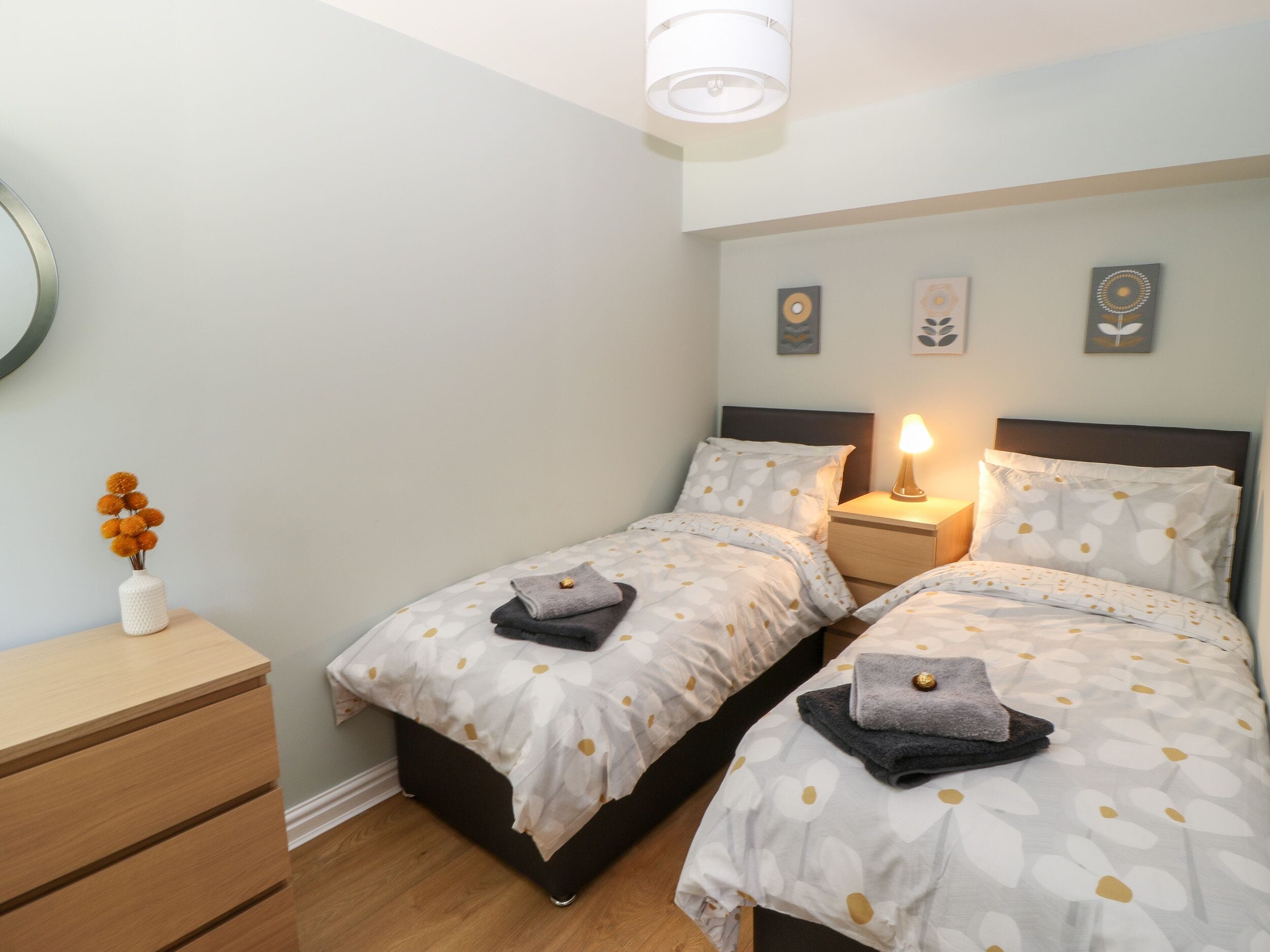 A bedroom with two beds, a bedside table with a lamp, a dresser, and wall art.