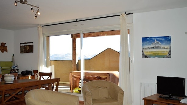 Ein gemütliches Wohnzimmer mit einer Couch, einem Sessel und einem Tisch mit einem Fernseher. Durch eine Glastür ist ein Balkon mit einem Holztisch und Stühlen zu sehen.