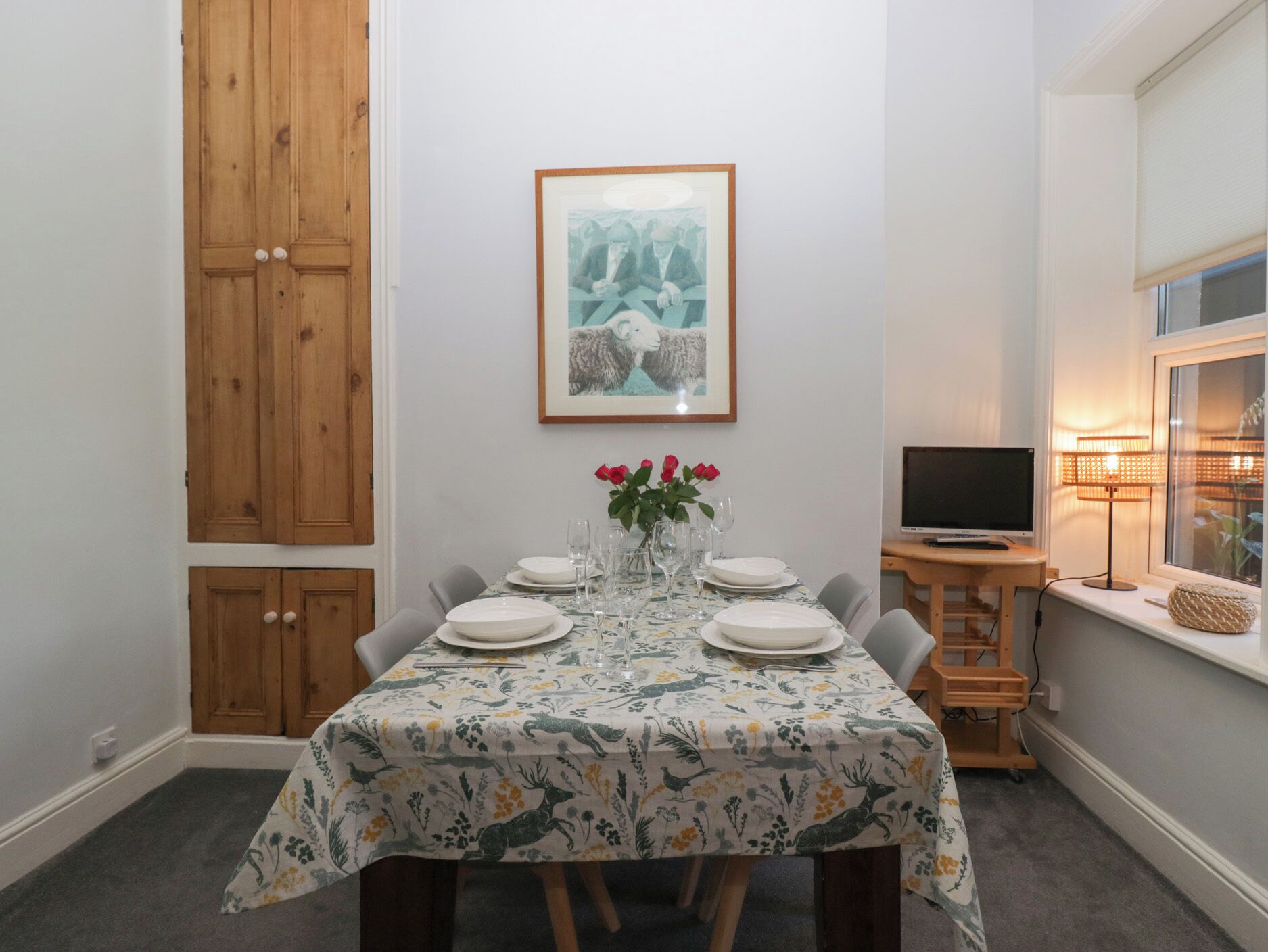 A dining area with a table set for six, a framed picture above the table, and a television on a stand to the right.