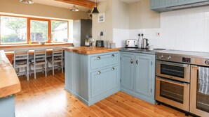 A kitchen with wooden flooring, a central island, light blue cabinets, stainless steel appliances, and a window with a view of greenery. - Ty Cerrig (Gaerwen)