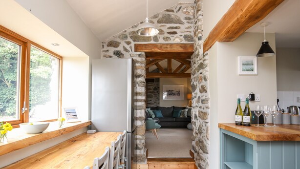 A kitchen with a stone wall, wooden beams, a dining area with chairs, a counter, and a view through to a living room. - Ty Cerrig (Gaerwen)