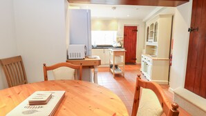 A dining area with a wooden table and chairs, a kitchen in the background, and a door leading to another room. - Compass Cottage (Holt)