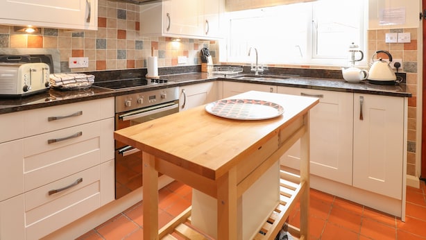 A modern kitchen with white cabinets, a wooden table, and a built-in oven. - Compass Cottage (Holt)