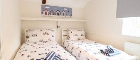 A room with two beds, each with a nautical-themed bedspread and pillows. There is a ceiling light and a wall-mounted shelf above the beds.