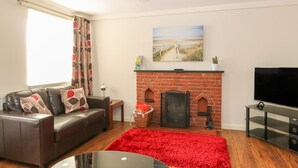 A living room with a leather sofa, a red rug, a fireplace, a TV, and a painting on the wall. - Compass Cottage (Holt)
