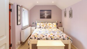 A bedroom with a bed, bedside tables, a nightstand, a window with blinds, a radiator, and a wall-mounted picture. - Compass Cottage (Holt)