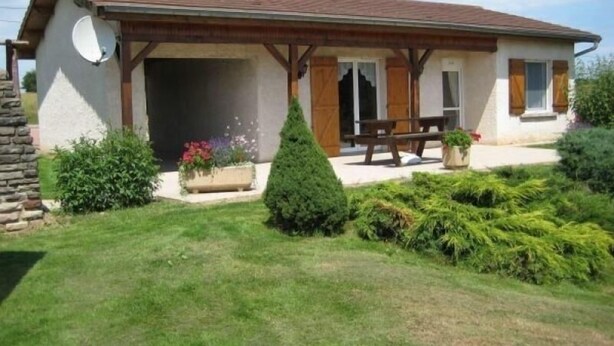 Ein einstöckiges Haus mit geneigtem Dach, einer Terrasse mit Tisch und Bänken sowie ein kleiner Garten mit verschiedenen Pflanzen und Blumen.