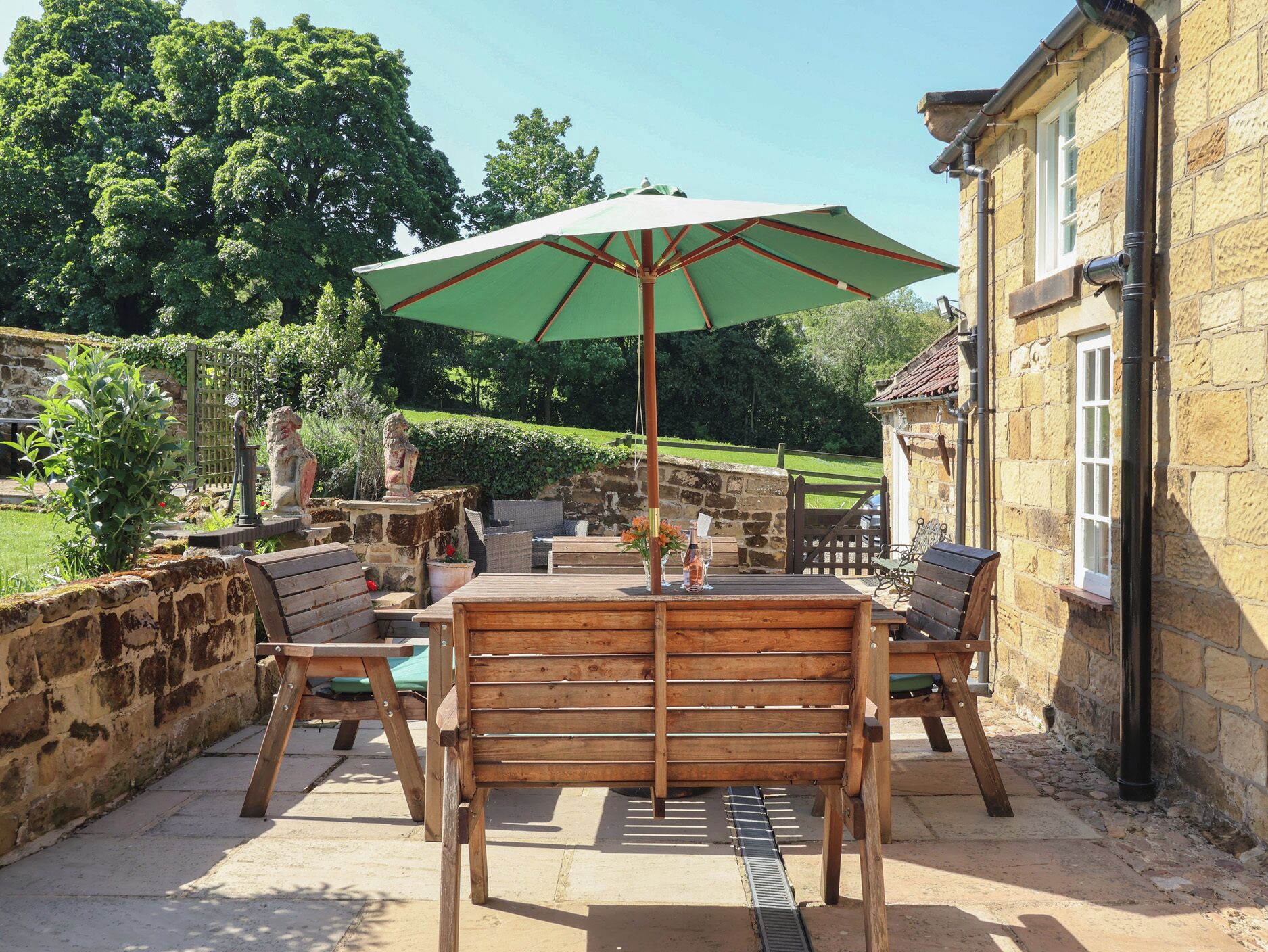 An outdoor patio with a wooden table and chairs, a green umbrella, and stone walls.