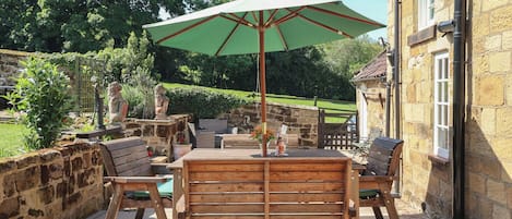 Een terras buiten met een houten tafel en stoelen, een groene parasol en stenen muren.