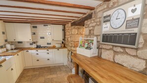Una cocina con armarios blancos, encimera de madera y pared de piedra. Hay un reloj y un calendario en la pared.