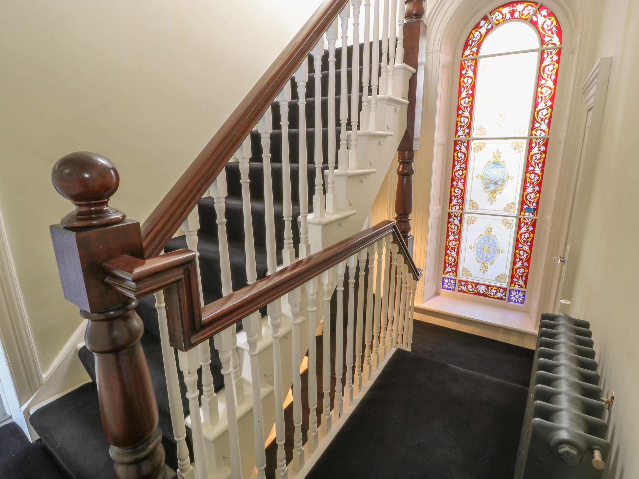 A staircase with a wooden handrail and white balusters, leading to a room with a black carpet and a radiator.
