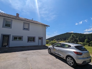 A white building with a tiled roof, a parked car with 'Gites de France' branding, and a forested hill in the background.