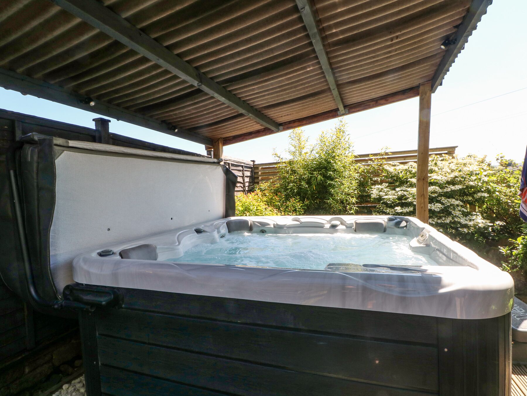 Un jacuzzi bajo un techo metálico, con un muelle de madera y follaje al fondo.