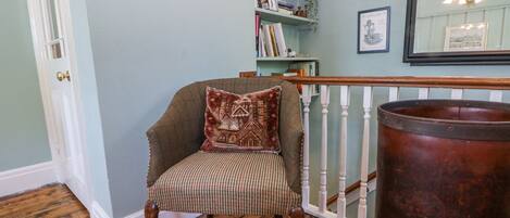 A room with a wooden floor, a chair with a patterned cushion, a bookshelf, and a large copper pot.