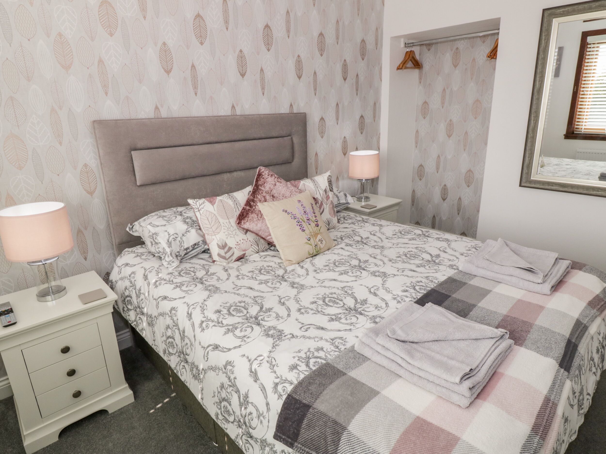 Una cama bien hecha con ropa de cama estampada, una lámpara de noche, una mesita de noche con un teléfono y un espejo en la pared.