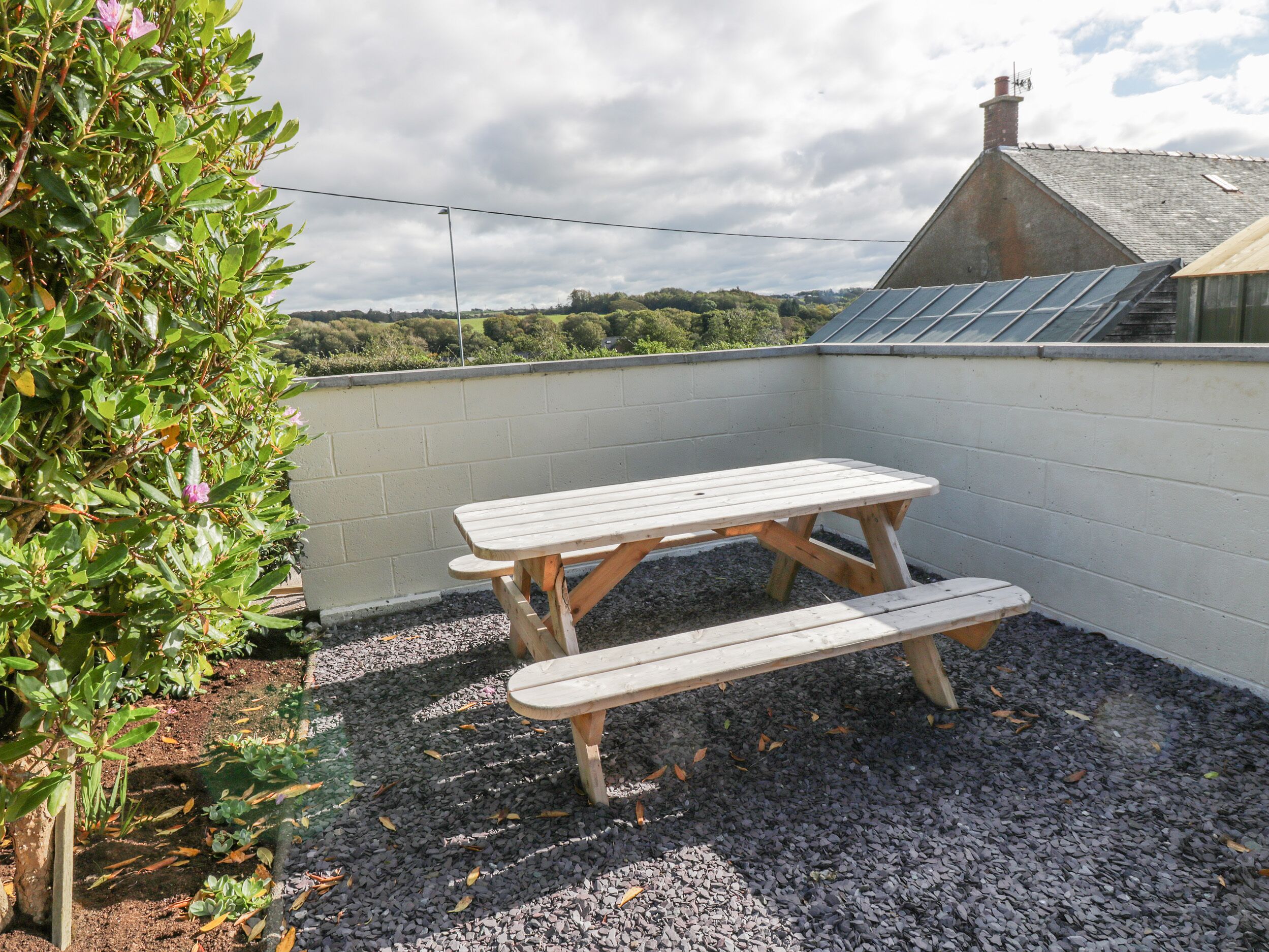 A wooden picnic table with benches in a garden area with gravel ground, surrounded by a white wall and a view of greenery and a building with a sloped roof.