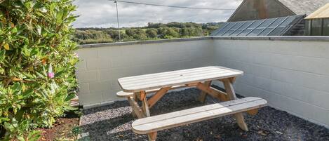A wooden picnic table with benches in a garden area with gravel ground, surrounded by a white wall and a view of greenery and a building with a sloped roof.