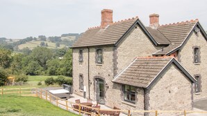 Un edificio de piedra con techo inclinado y chimeneas, que cuenta con un patio con mobiliario exterior y vistas al campo.