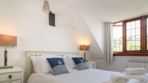 Una cama bien hecha con ropa blanca y almohadas azul marino, una mesita de noche con lámpara y una ventana con cortinas.