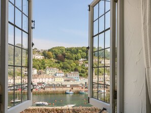 A view through open windows of a coastal town with buildings, a harbor, and a hill with trees. - Pixie Cottage (Looe)