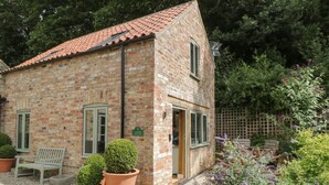 Une petite maison en briques avec un toit en tuiles, un banc, des plantes en pot et un jardin où poussent des fleurs violettes.