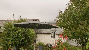 A garden with a round table and an umbrella, surrounded by lush greenery and a house with a white exterior and a tiled roof. - The Old Bakery (Taunton)