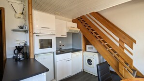 Una cucina compatta con mobili bianchi, una macchina da caffè, un forno a microonde e una lavatrice. Una scala in legno conduce a un piano superiore.