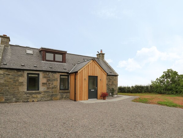 A stone house with a wooden extension, a gravel driveway, and a garden. - Enzie Station Cottage (Buckie)