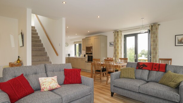 A modern living room with two grey sofas, a wooden staircase, a kitchen, and a dining area. - Enzie Station Cottage (Buckie)