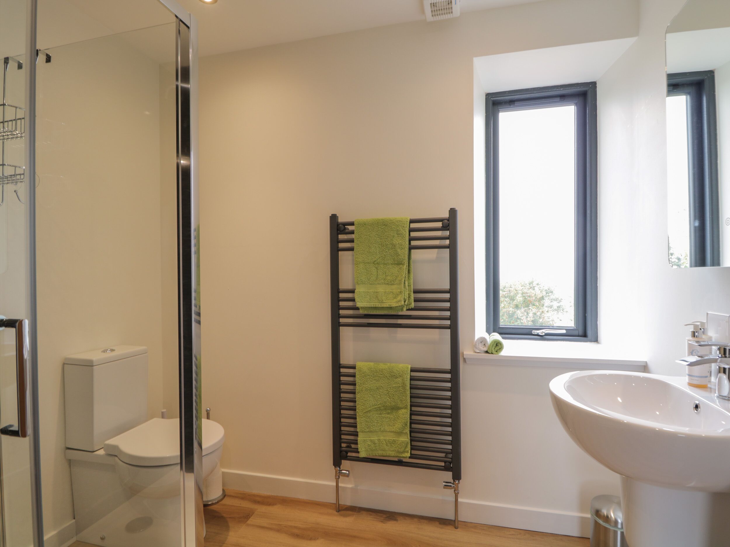 Un baño con cabina de ducha de vidrio, inodoro blanco, toallero negro con toallas verdes, ventana con vistas y lavabo blanco.