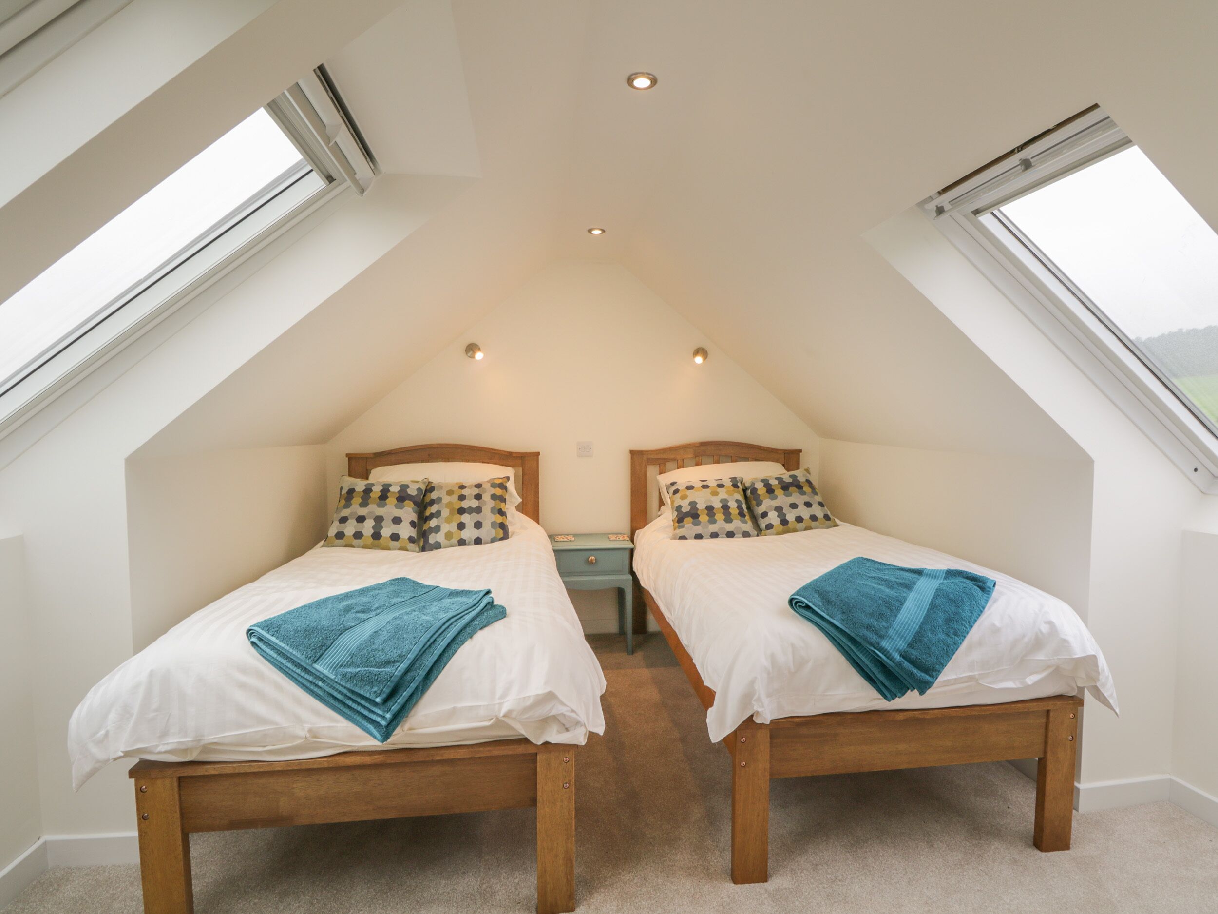 A bedroom with two beds, each with a wooden frame and white bedding, under a sloped ceiling with skylights.