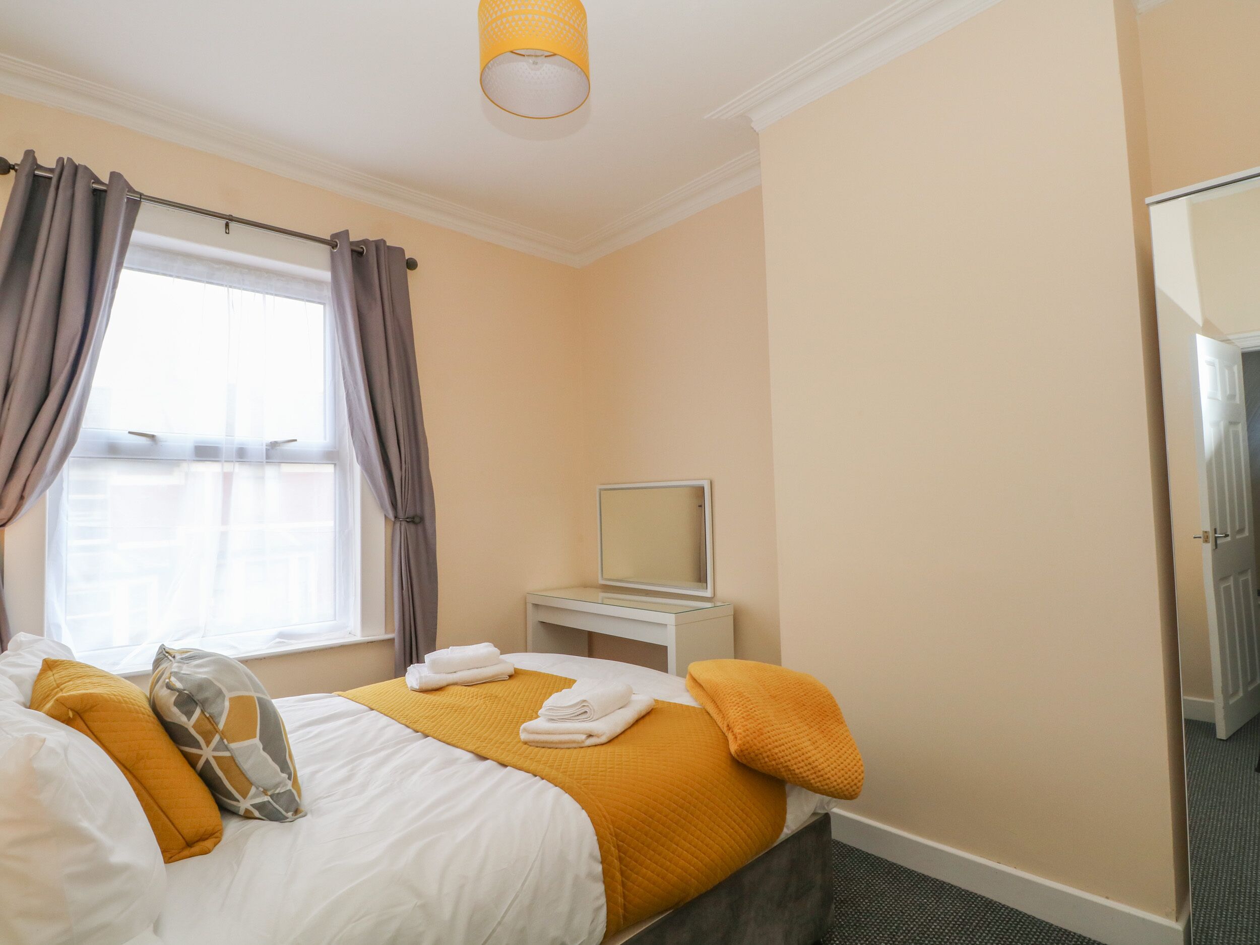 A hotel room with a bed, a bedside table with a mirror, a window with curtains, and a door.