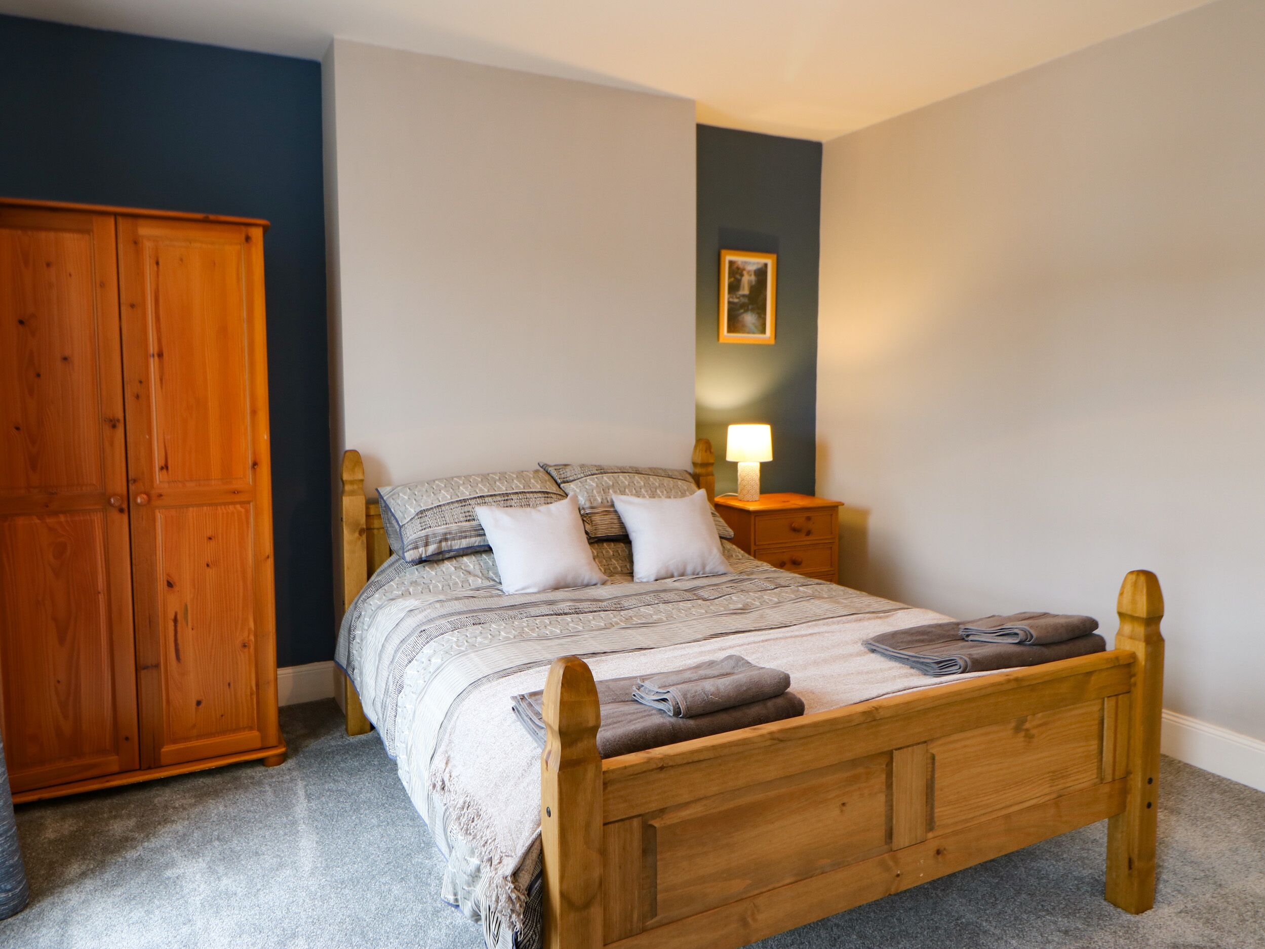 A bedroom with a wooden bed, a nightstand with a lamp, a wardrobe, and a framed picture on the wall.