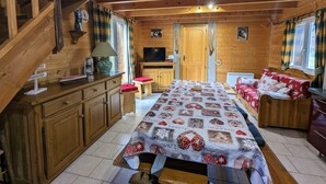 A cozy cabin interior with a wooden staircase, a dining table with a patterned tablecloth, a sofa with red cushions, and a television on a wooden cabinet. - Cozy Chalet by the Stream: Terrace, BBQ, Near Skiing & Cycling Paths, Pet Friendly (Vagney)