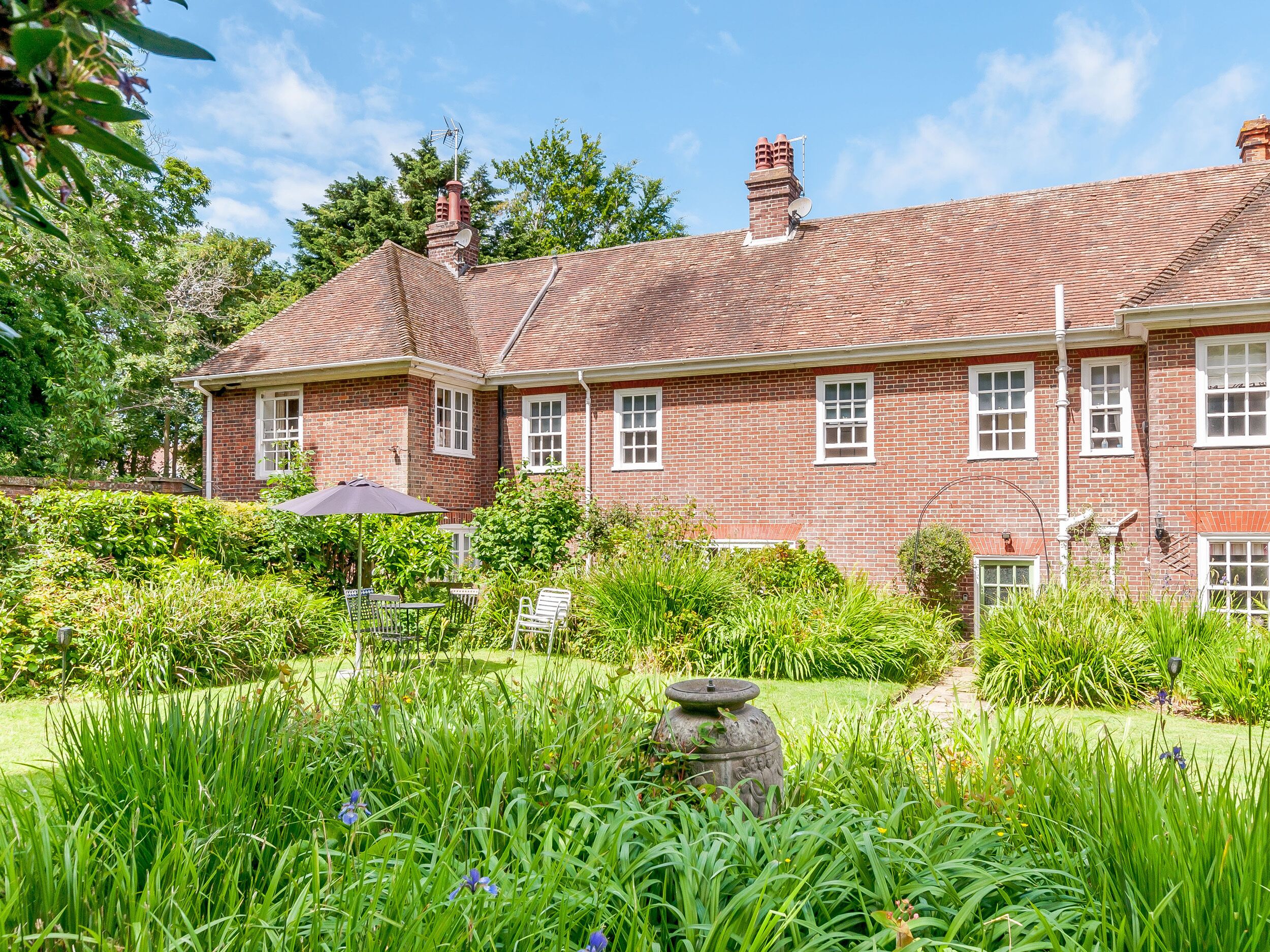 A large brick house with a tiled roof, surrounded by lush greenery and a garden with a stone fountain.