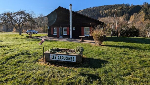Une cabane en bois rustique, dotée d’une cheminée, sur laquelle on peut lire l’inscription « LES CAPUCINES », et un champ verdoyant au premier plan.