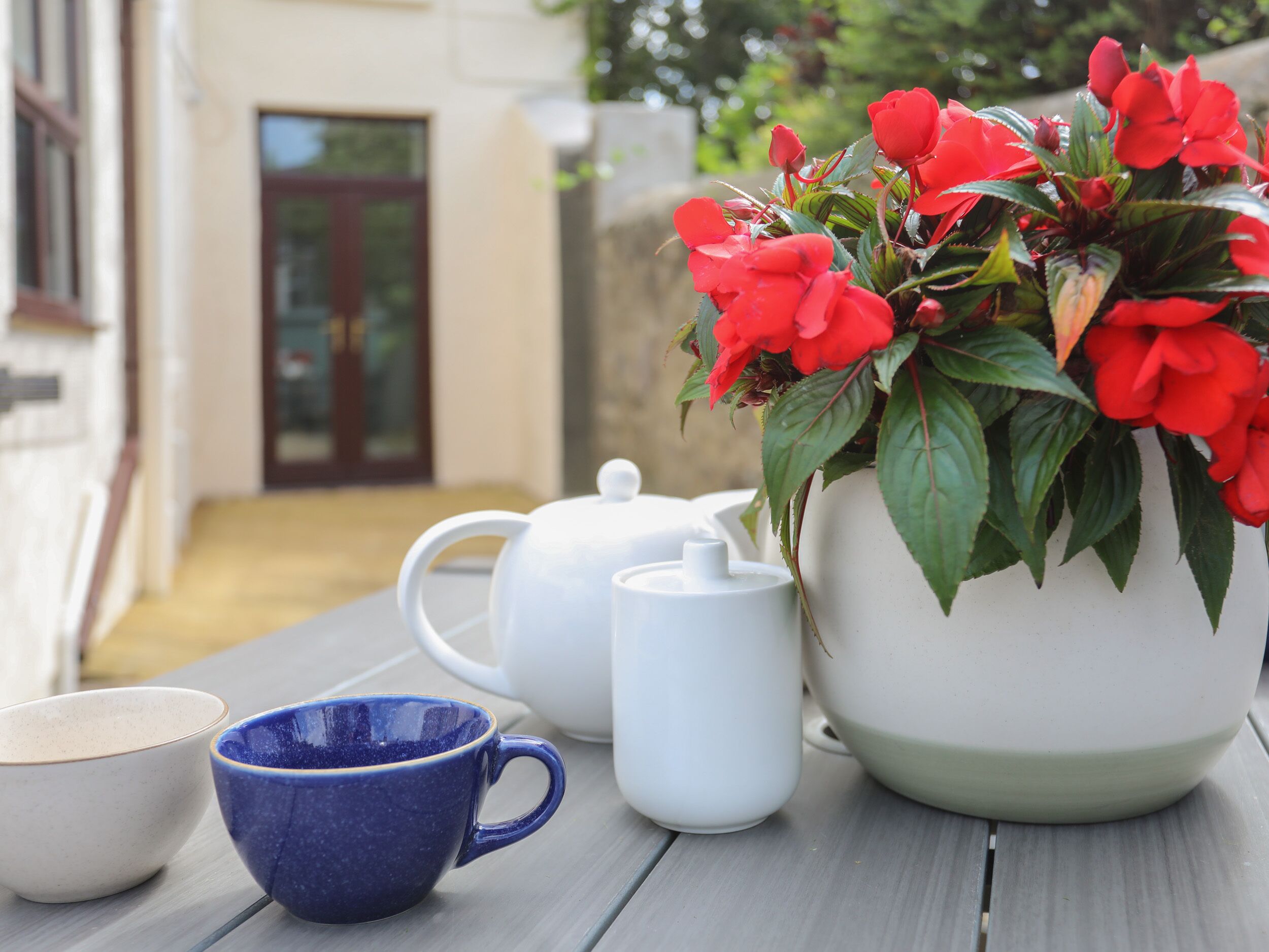 Une table sur laquelle se trouvent une théière blanche, une tasse bleue et une soucoupe blanche, ainsi qu’une plante en pot ornée de fleurs rouges.