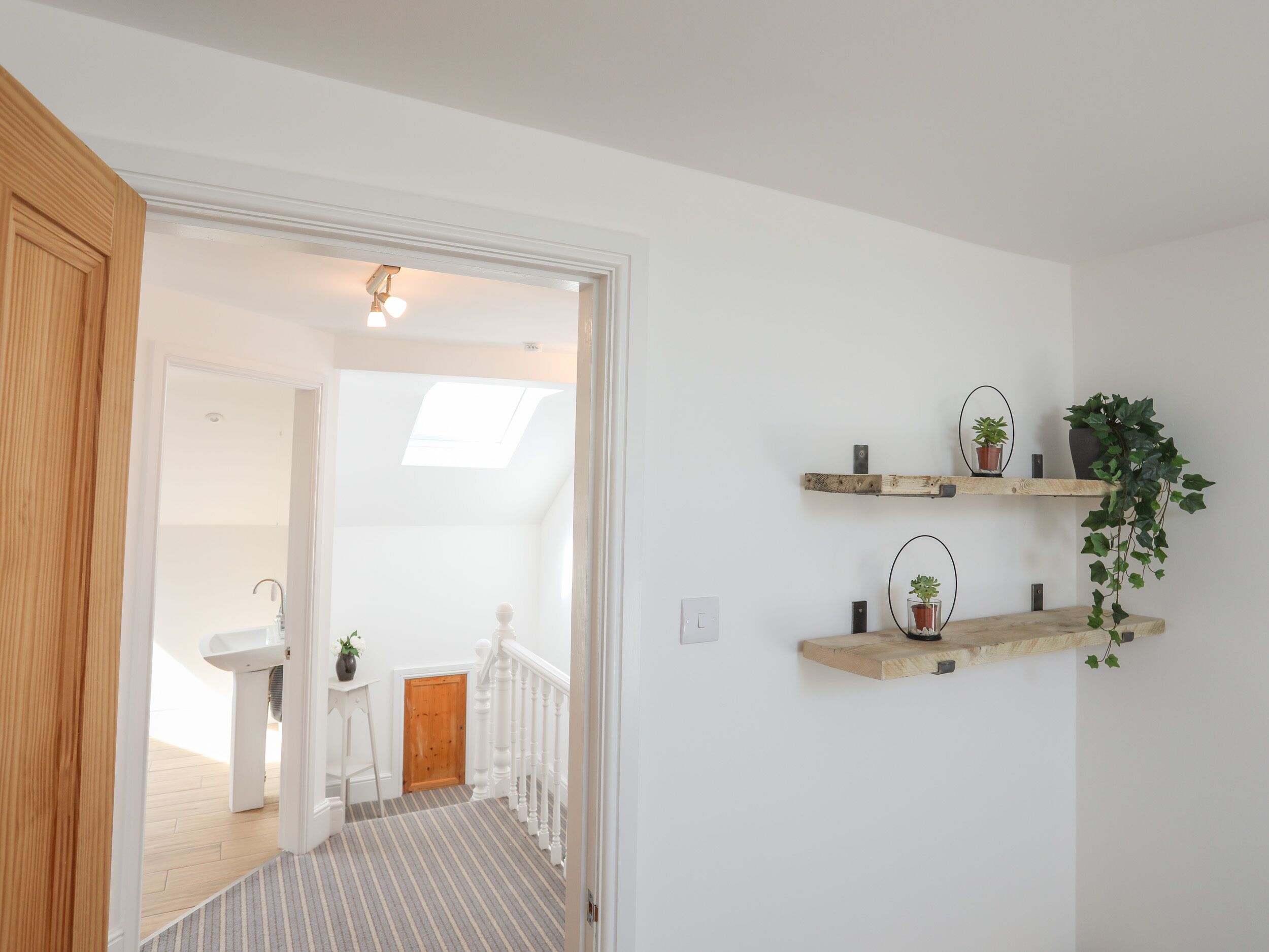 A hallway with a wooden door, a white wall, and a staircase with a striped carpet. There are two wooden shelves with plants and a small table with a vase.