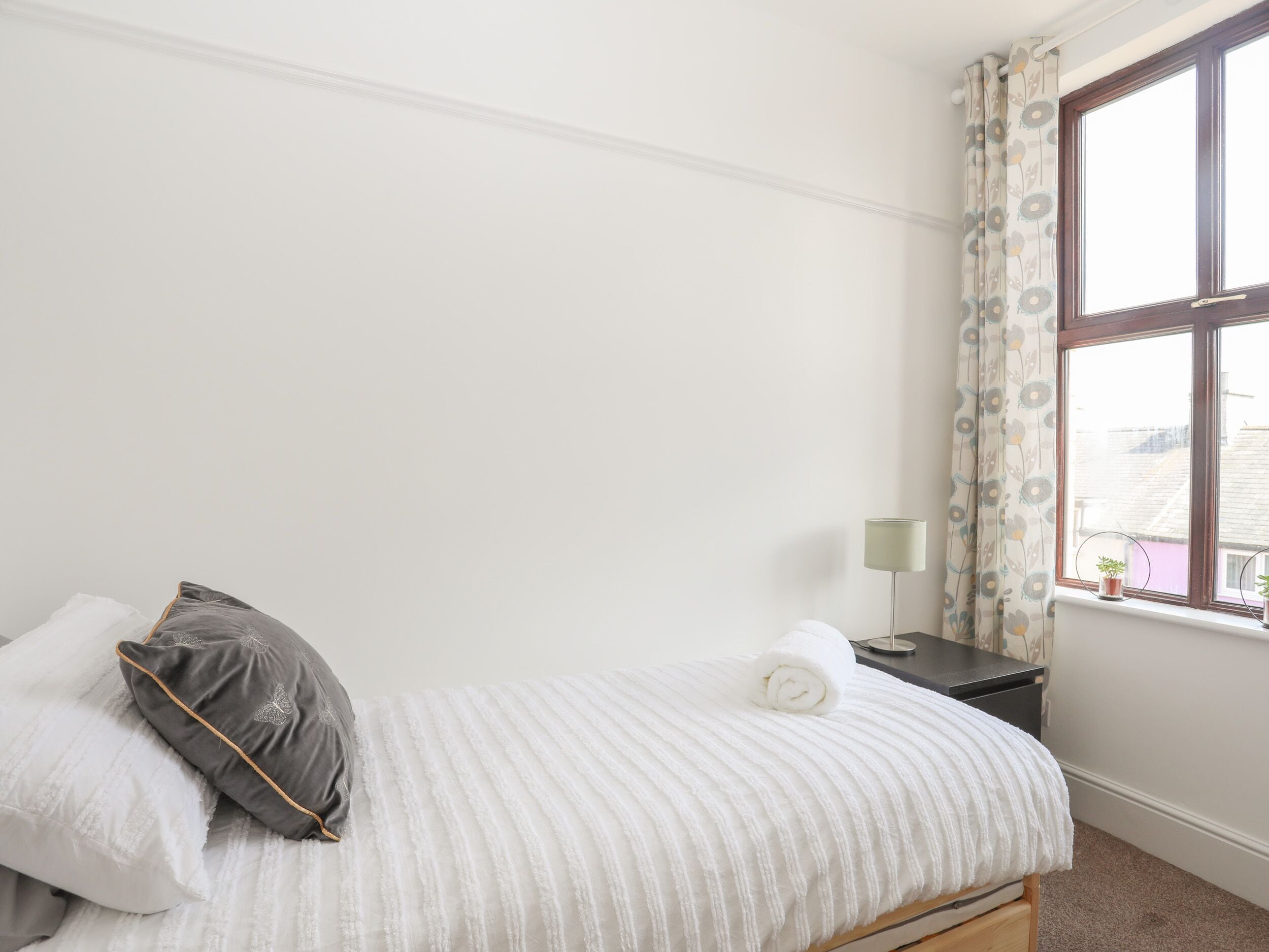 Cama bem arrumada com travesseiros, mesa de cabeceira com abajur e janela com cortinas.