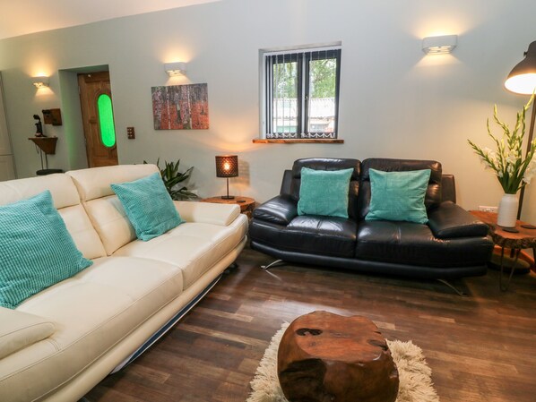 A living room with a beige sofa, a black leather couch, and a wooden coffee table. There are two wall-mounted lamps, a painting, and a window with blinds.