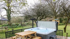 An outdoor hot tub with a wooden table and benches, surrounded by a garden and trees. - Elvan Cottage (Liskeard)