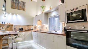 A modern kitchen with white cabinets, a built-in oven, and a microwave. There is a sink with a faucet, a countertop with a kettle, and a plant on the window sill. - Elvan Cottage (Liskeard)