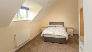 A bedroom with a sloped ceiling, a bed with a patterned bedspread, a nightstand, a dresser, and a window with a view of greenery outside. - Woodpecker Barn (Pontyclun)