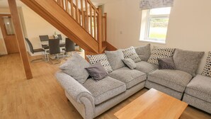 A living room with a wooden staircase, a grey sectional sofa with patterned cushions, a wooden coffee table, and a dining area in the background. - Woodpecker Barn (Pontyclun)