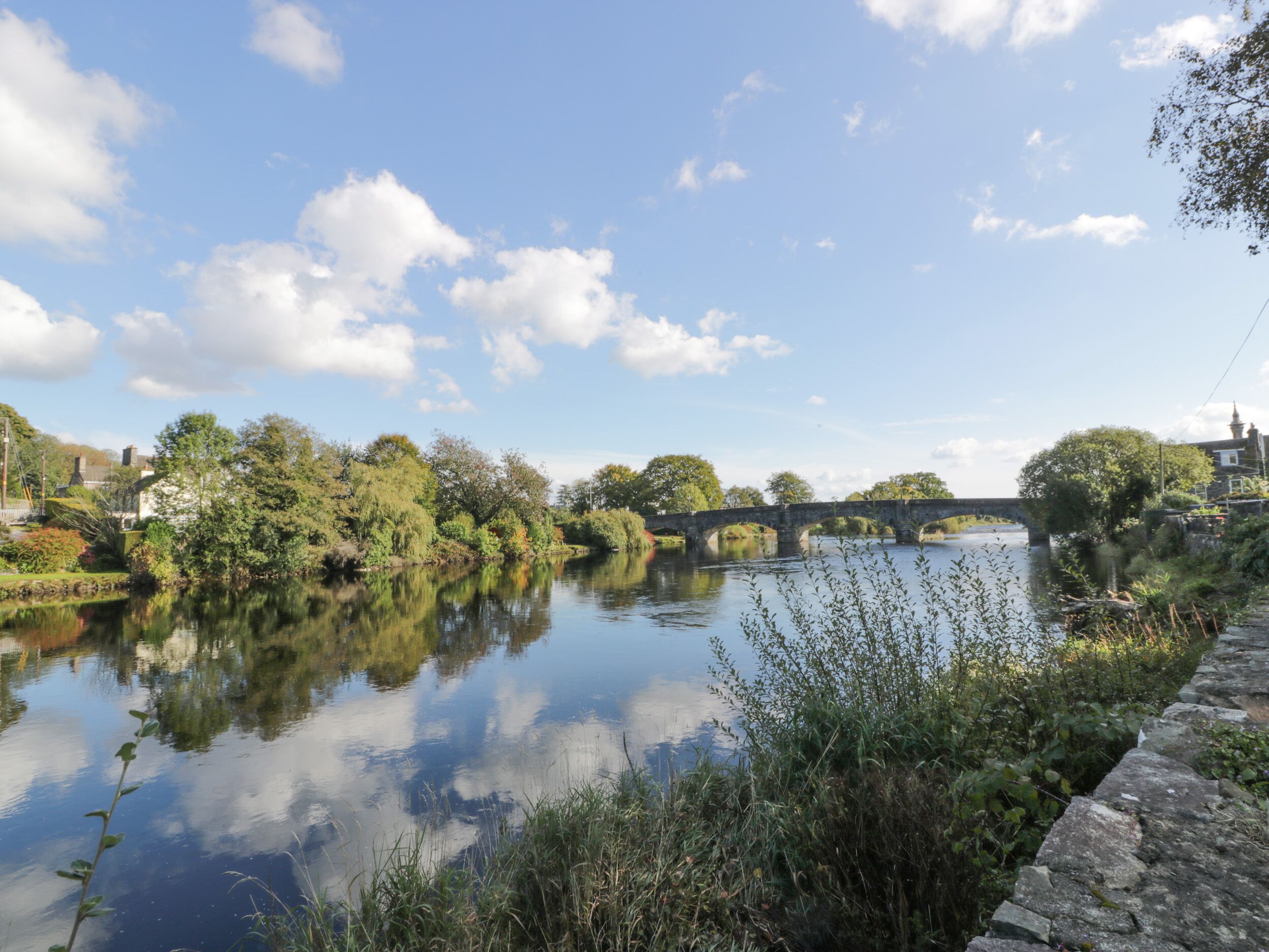 Un pont de pierre enjambe une rivière paisible, bordée de chaque côté d’une végétation luxuriante et d’arbres.