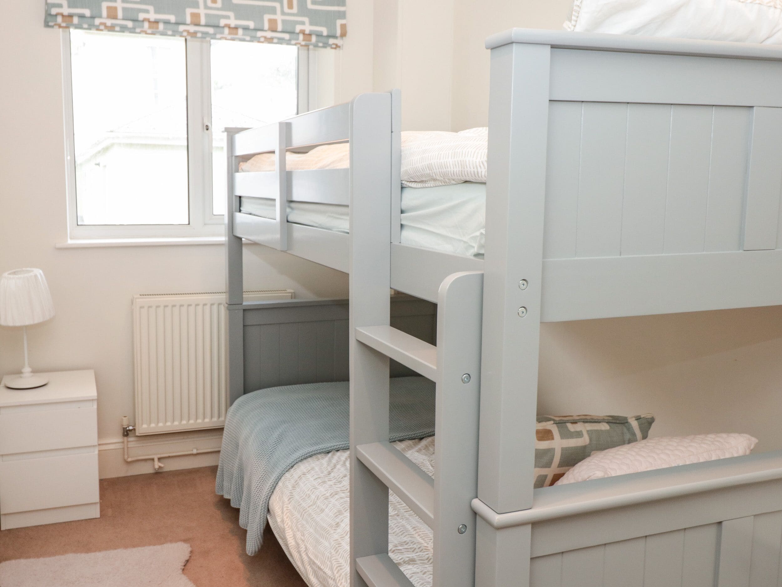 A bunk bed room with a white nightstand and a lamp, a radiator, and a window with curtains.