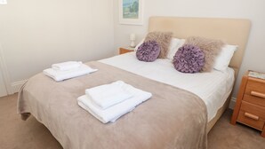 A neatly made bed with two folded white towels, a beige bedspread, and two decorative pillows on a headboard.