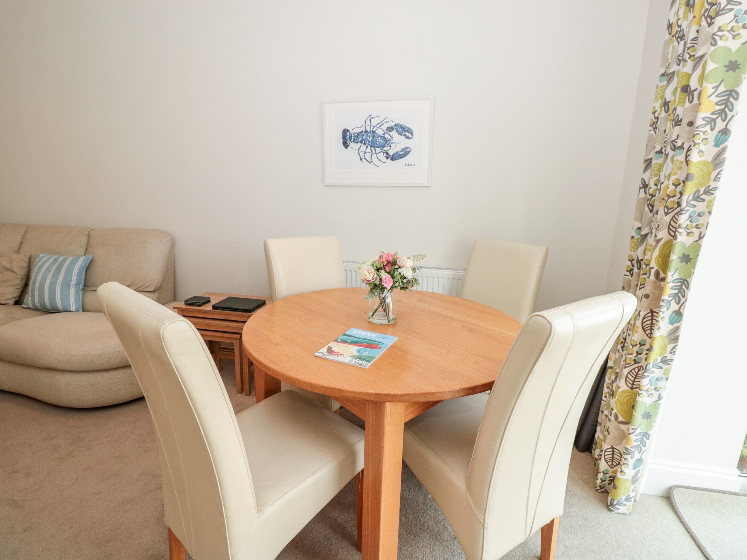 A cozy living room with a round wooden table, four beige chairs, a beige sofa, a framed artwork on the wall, and a floral curtain.