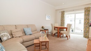 A living room with a beige sofa, a wooden coffee table, and a dining table with four chairs. There is a window with curtains, a radiator, and a small table with a vase of flowers.