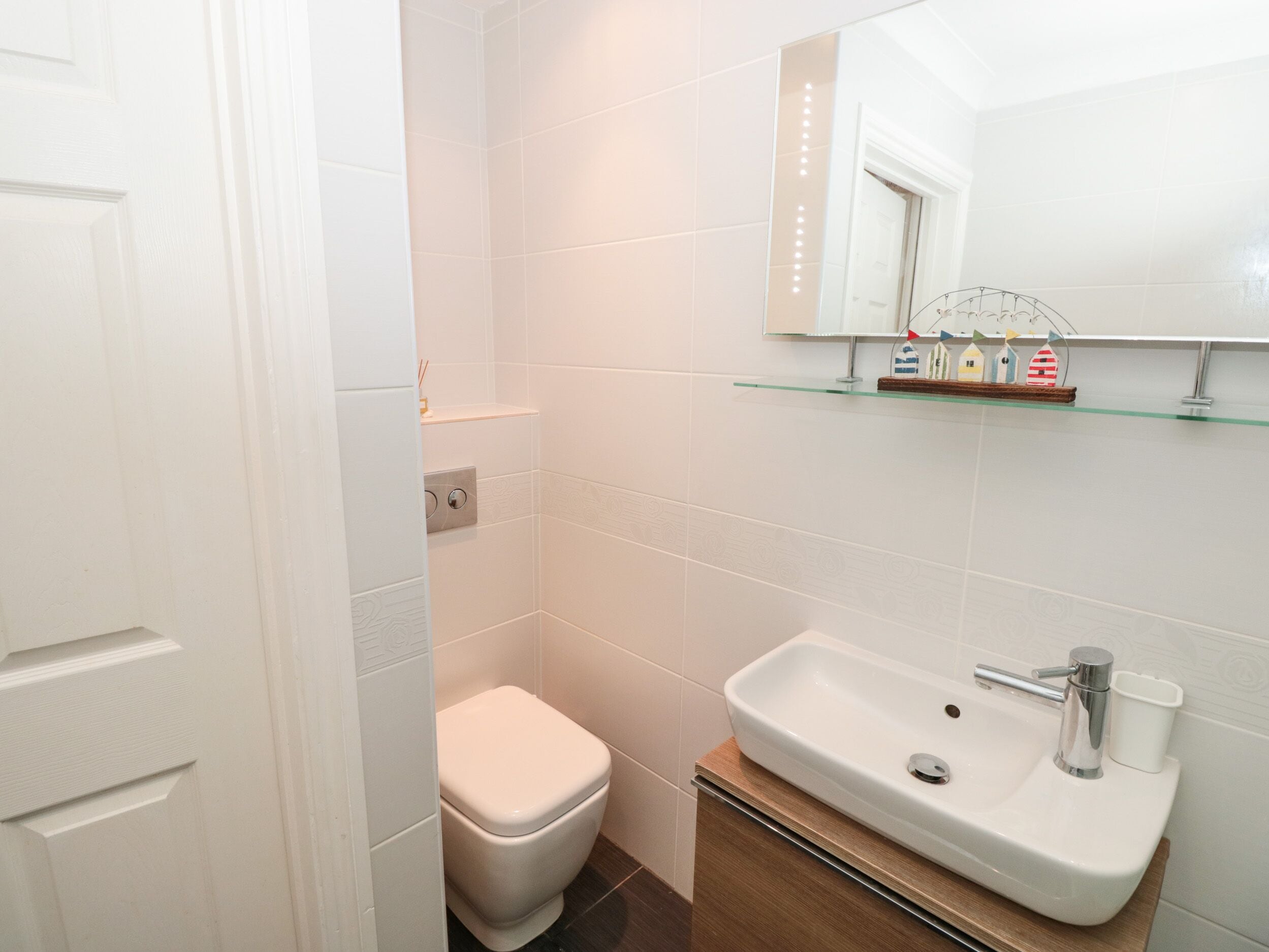 A bathroom with white tiles, a white toilet, a sink with a mirror above it, and a shelf with decorative items.